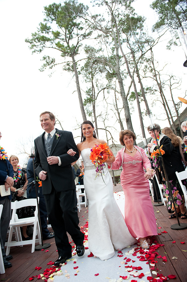 bride being escorted down aisle by mother and father - bride is wearing white a-line dress and holding orange, coral, and dark pink bouquet, father is wearing black suit with gray vest and tie and mother is wearing light pink dress - photo by Houston based wedding photographer Adam Nyholt